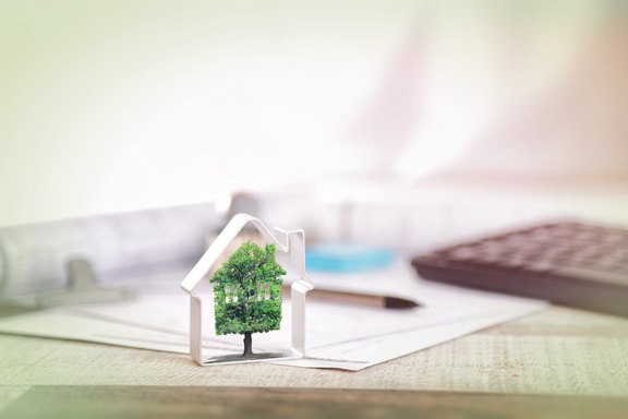 Ökologisch Bauen Ein kleine Baum in einem Hausmodell integriert, steht auf einem Tisch, auf dem auch Unterlagen liegen